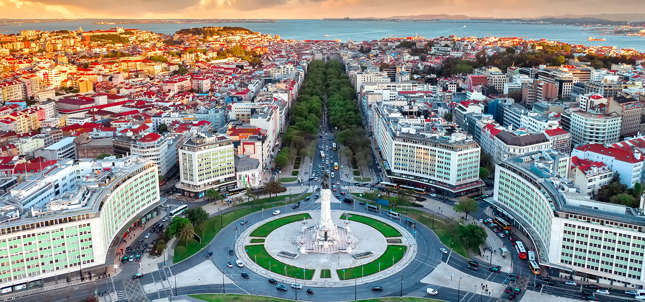 Aerial view of Lisbon at sunset with a monument roundabout, a tree-lined avenue and the river beyond